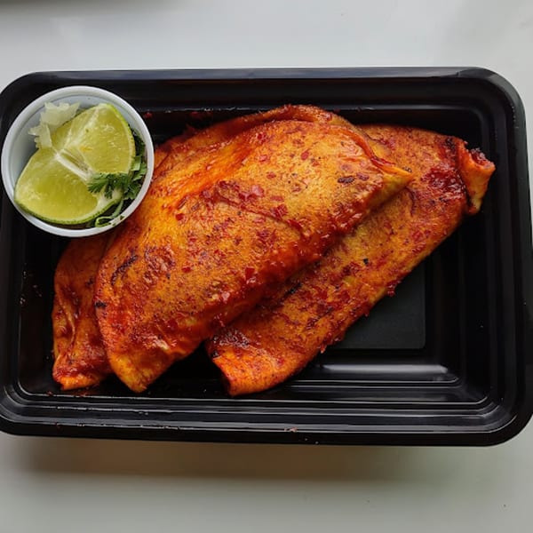 Three red-orange Mexican empanadas in a black to-go container with a side of lime wedges and cilantro.