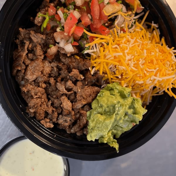 A black bowl filled with Mexican food, including steak, pico de gallo, shredded cheese, and guacamole.